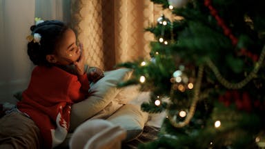 Child lying by a decorated Christmas tree, gazing with wonder and holiday joy indoors.