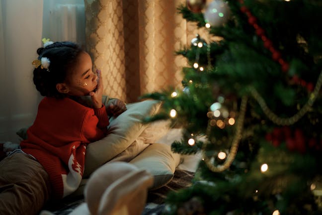 Child lying by a decorated Christmas tree, gazing with wonder and holiday joy indoors.